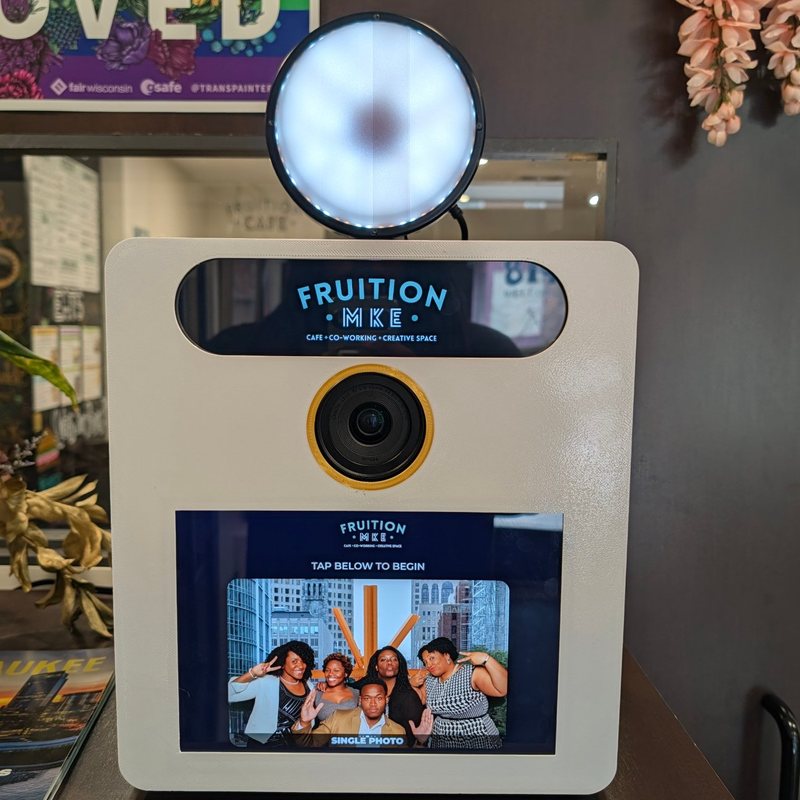 The Booth in white with a gold lens ring and the Fruition MKE logo on the billboard